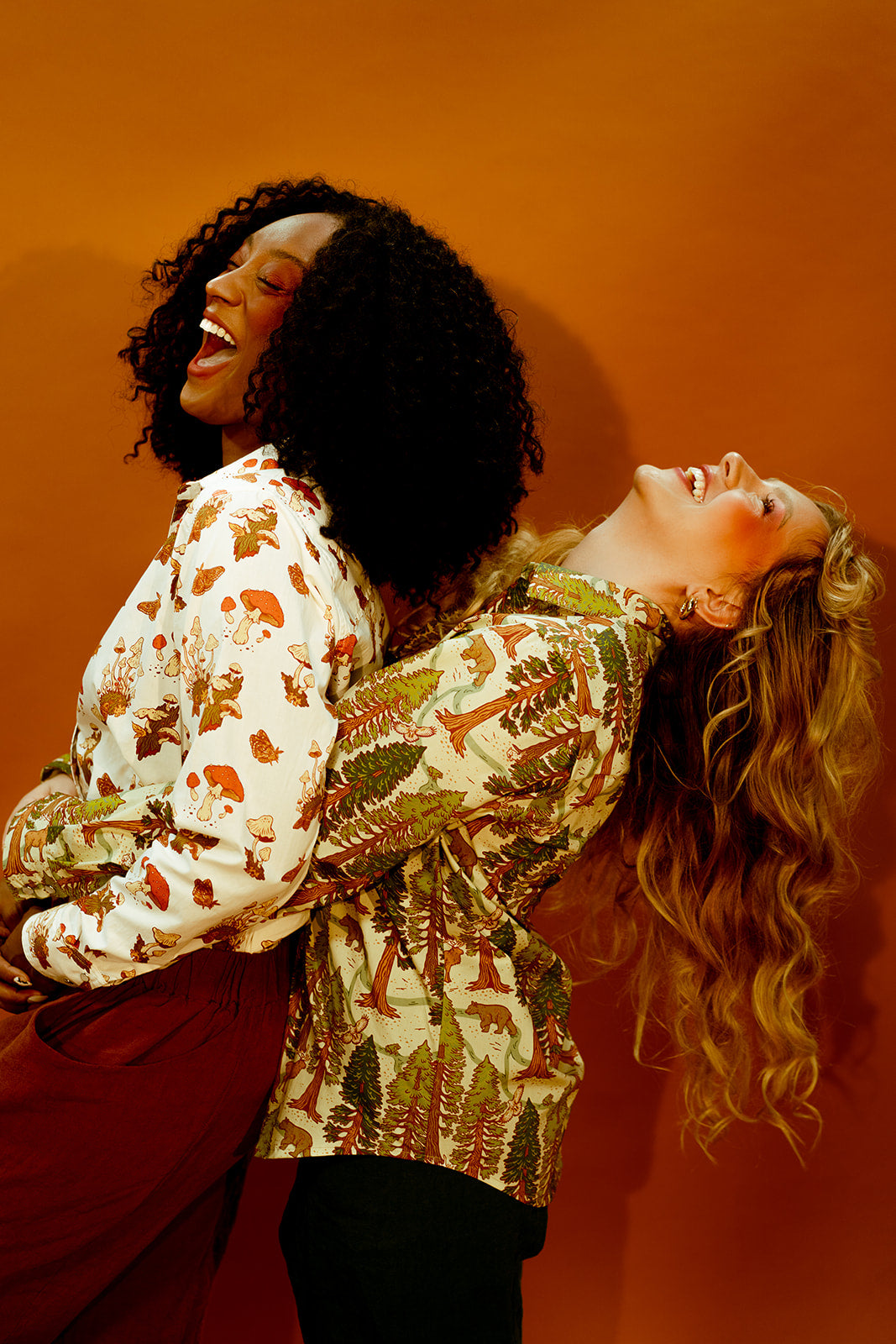 Two happy women, one in a mushroom-print Western-style shirt with red pants and the other in a California Redwoods-print shirt with black pants, stand together. The back woman hugs the other from behind, against a dark brown background.