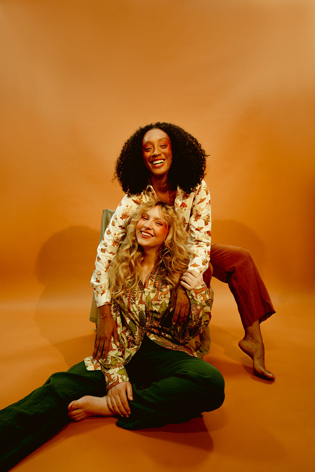 A couple of white and black women with beautiful smiles are sitting and posing for a picture. A white woman is sitting on the floor, wearing an aesthetically groovy Western-style front snap shirt cast in the beautiful greens and tans of California Redwoods. Meanwhile, a black woman wearing a Western-style front snap shirt in a groovy mushroom print sits on top of her against a brown wall.