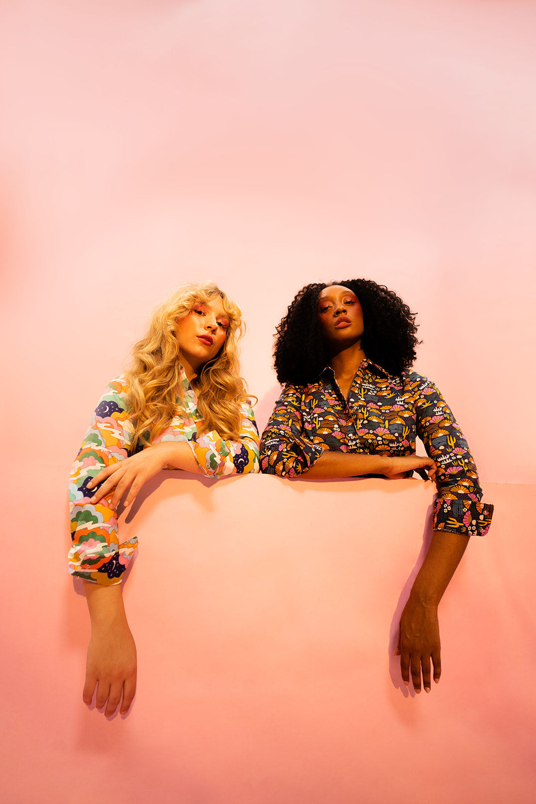 Two Bold women are sitting side by side on a pink background, wearing pearl snap shirts with mushroom and redwoods prints.