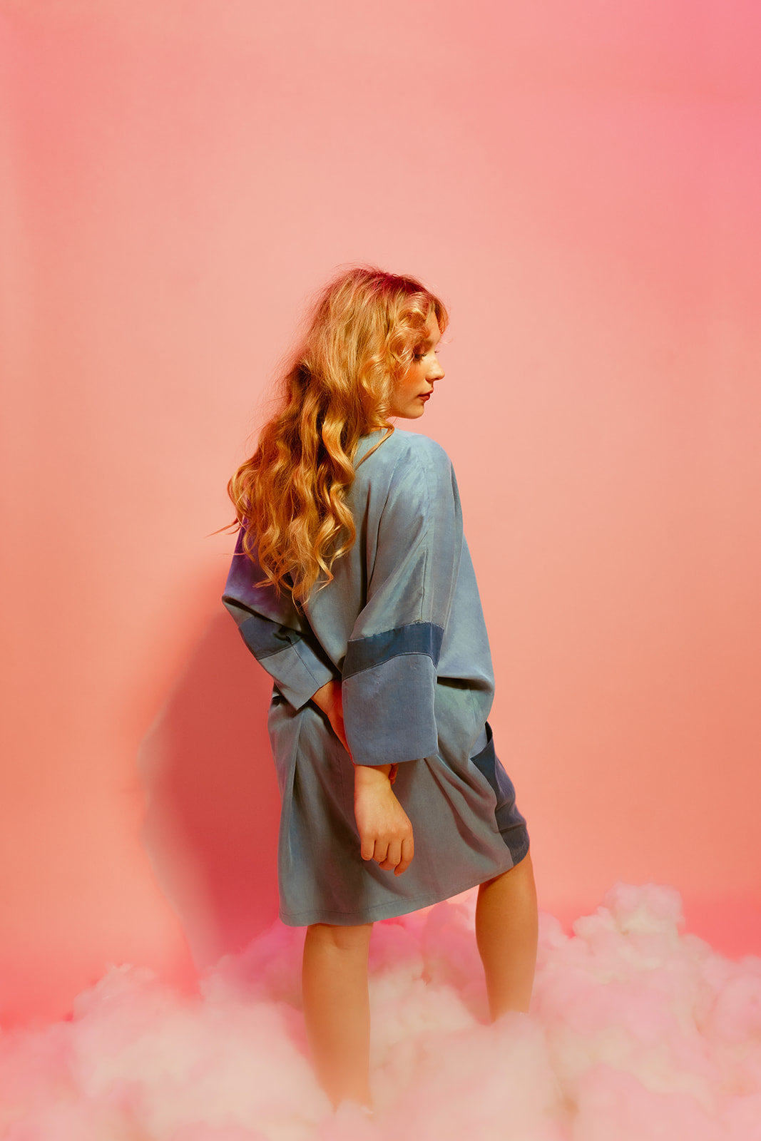 A woman is standing on clouds, posing for a picture in front of a pink background. She is wearing a blue gown dress.